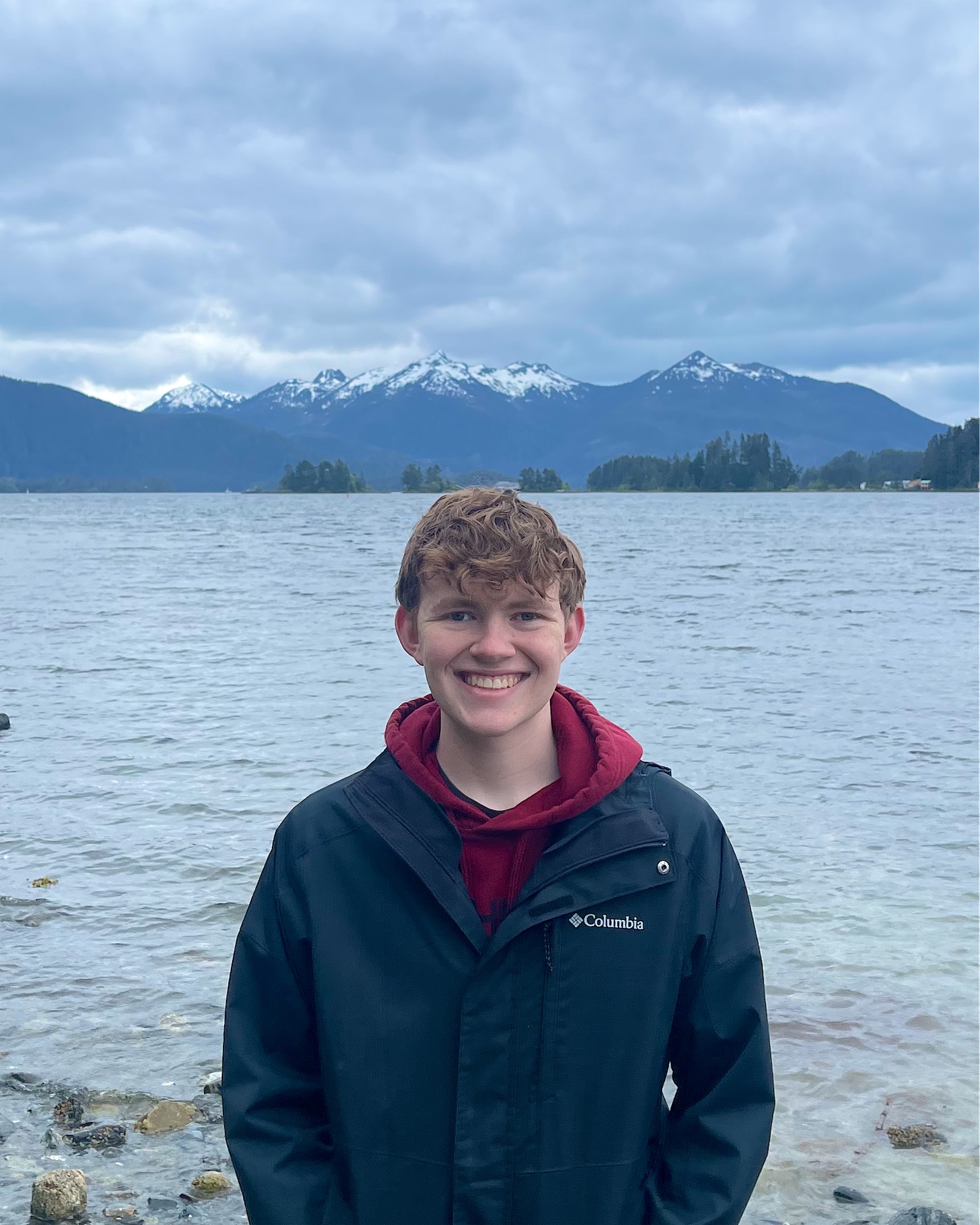 Eric smiling on a rocky beach with snow-capped mountains behind him
