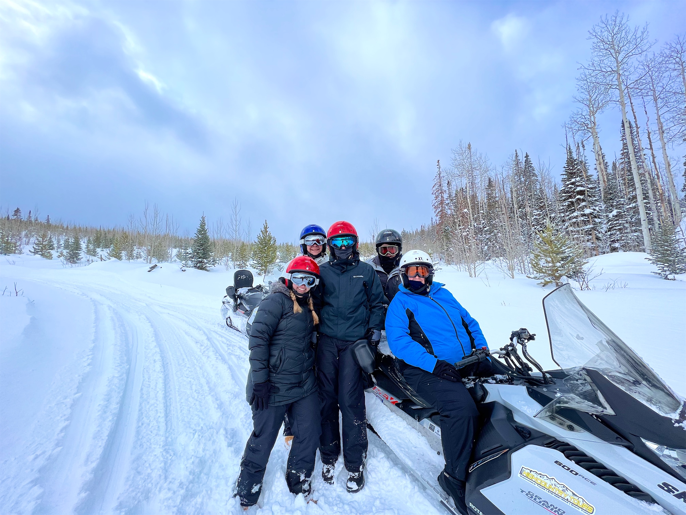 Four people in helmets and goggles posing with snowmobiles on a snowy trail